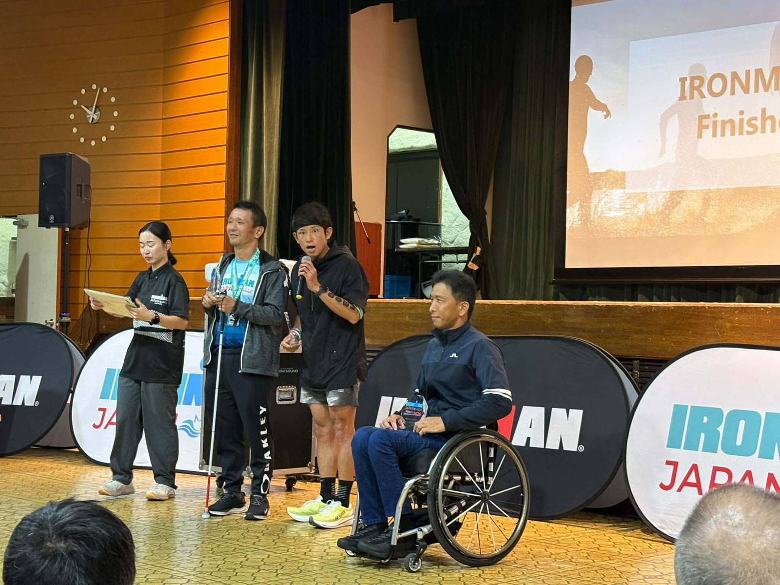 IRONMAN JAPAN 2026の表彰式で、白杖を持ちフィニッシャートロフィーを手にする中澤隆